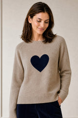Model wearing the Aspiga Cashmere Crew Neck Heart Jumper in Oat/Navy, a crew-neck jumper with a contrast heart motif, over navy trousers, front view