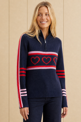 Model wearing the Aspiga Merino Wool Stripe Hearts Quarter Zip in Navy/Red/Light Pink with navy trousers, front view, smiling