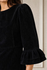 Model wearing the Aspiga Elizabeth Stretch Corduroy Midi Dress in Black with strappy heels, full-length front view, smiling