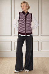 Model wearing the Aspiga Merino Wool Stripe Zip Through Gilet in Burgundy/Pink over a white blouse with black trousers, full-length front view, hands in pockets