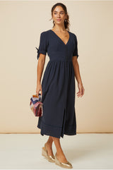 Woman wearing a navy midi dress with short tie sleeves, button front detail, and a front slit, styled with gold wedge espadrilles and a multicoloured handbag.
