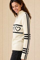 Model wearing the Aspiga Merino Wool Stripe Hearts Quarter Zip in Off White/Black with black jeans, side view, smiling, looking down