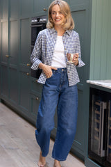 Model wearing the Aspiga Classic Gingham Shirt in Blue/White with jeans, smiling, holding a teacup in a kitchen
