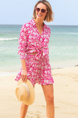 Woman walking on a beach wearing a bright pink and white floral smocked mini skirt with ruffle hem, paired with a matching tied shirt.