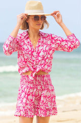 Woman on a beach wearing a vibrant pink and white floral smocked mini skirt with a ruffle hem, paired with a matching tied shirt, sunglasses, and a straw hat.