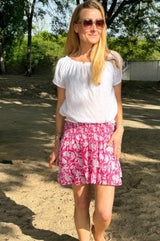 Woman standing outdoors wearing a pink and white floral smocked mini skirt with a ruffle hem, paired with a white short-sleeve blouse and sunglasses.