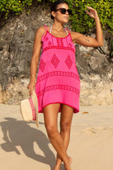 A woman wearing a pink and red embroidered cotton dress with a scoop neckline and spaghetti tie straps, standing on a beach holding Aspiga's Panama hat
