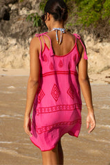 A woman wearing a pink and red embroidered cotton dress with a scoop back and spaghetti tie straps, standing on a beach with a bikini underneath