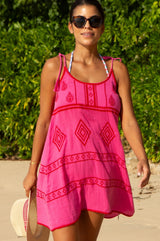 A woman wearing a pink and red embroidered cotton dress with a scoop neckline and spaghetti tie straps, standing on a beach.
