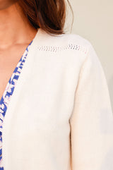 Close-up of a cream knit cardigan with delicate openwork detailing along the shoulder seam, worn over a blue floral dress.