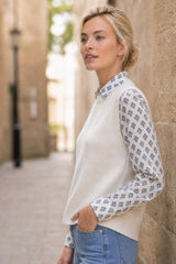 Model wearing the Aspiga Merino Wool Knitted Short Tank in Ivory layered over a printed blouse with jeans, side view leaning against a stone wall
