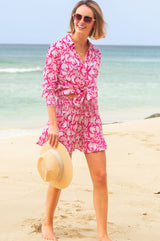 Woman wearing a pink and white floral print long-sleeve shirt tied at the waist, styled with matching shorts, sunglasses, and a straw hat on the beach.