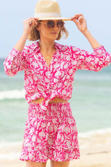 Woman wearing a pink and white floral print long-sleeve shirt tied at the waist, styled with matching shorts, sunglasses, and a straw hat on the beach.