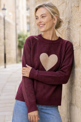 Model wearing the Aspiga Cashmere Crew Neck Heart Jumper in Burgundy/Oat, leaning against a stone wall outdoors, smiling