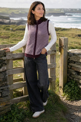 Model wearing the Aspiga Merino Wool Stripe Zip Through Gilet in Burgundy/Pink, standing by a wooden gate outdoors by the coast