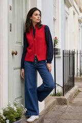Model wearing the Aspiga Merino Wool Zip Through Gilet in Red over a navy lace top with jeans, leaning against a door, one leg raised