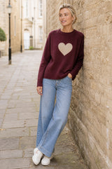 Model wearing the Aspiga Cashmere Crew Neck Heart Jumper in Burgundy/Oat with jeans, walking outdoors and leaning against a stone wall