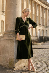 Model wearing the Aspiga Lorna Lace Velvet Dress in Dark Olive with a gold clutch, leaning against a stone column outdoors, smiling