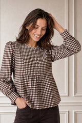 Model wearing the Aspiga Mina Gingham Check Blouse in Chocolate/White, a peplum blouse with puff sleeves and scalloped collar, hand in hair, smiling