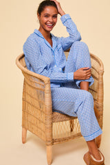 Woman wearing a light blue button-up pyjama set with a small navy floral print and contrast piping, paired with tan shearling slippers.