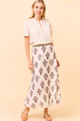 Woman wearing a white maxi skirt with pink and green floral block print, paired with a delicate short-sleeve cream blouse and nude Mary Jane flats.