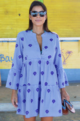 Woman wearing a light blue embroidered tiered dress with long sleeves and sunglasses, holding a pair of sandals.