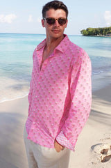 A man wearing sunglasses and a periwinkle pink/rust colored long-sleeved cotton shirt with a classic collar paired with neutral linen trousers on a beach