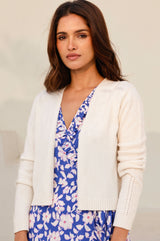 Woman wearing a cropped cream knit cardigan with subtle openwork details on the shoulders and sleeves, styled over a blue floral dress.