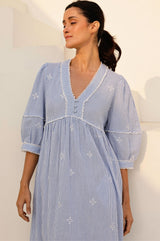 Woman wearing a light blue and white striped midi dress with embroidered details, pom-pom trim along the neckline and cuffs, and 3/4 puff sleeves.