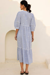 Back view of a light blue and white striped midi dress with white embroidery, pom-pom trim, and tiered skirt, featuring three-quarter sleeves and a relaxed fit.