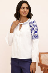 Woman wearing a white long-sleeve blouse with blue floral embroidery on the shoulders and sleeves, and navy trousers.