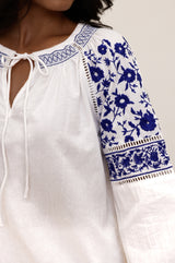Close-up of a white blouse with blue floral embroidery on the shoulders and sleeves, featuring a tie neckline and decorative stitching.