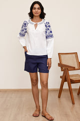 Woman wearing a white blouse with blue floral embroidery on the shoulders and sleeves, featuring a tie neckline and long sleeves, styled with navy shorts and tan sandals.