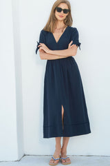 Woman wearing a navy midi dress with short sleeves tied at the cuffs, a buttoned bodice, a v-neckline, and a front slit, paired with patterned sandals and sunglasses.