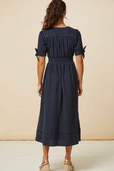 Back view of a woman wearing a navy midi dress with short tie sleeves, styled with gold wedge espadrilles.