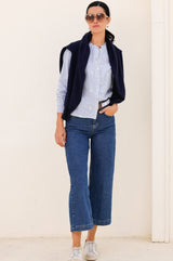 A woman wearing a blue and white long-sleeved Cosima organic cotton shirt with embroidered motifs and shirred elasticated cuffs styled with wide leg cropped denim Aspiga jeans and a navy knit jumper over her shoulders