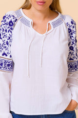 Close-up of a white blouse with blue floral embroidery on the shoulders and sleeves, featuring a tie neckline and long sleeves.