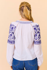 Back view of a woman wearing a white blouse with blue floral embroidery on the shoulders and sleeves, paired with blue jeans.