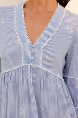 Close-up of the bodice of a blue and white striped dress with delicate embroidery, fabric-covered buttons, and white pom-pom trim along the V-neckline and seams.