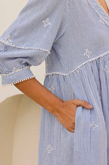 Close-up of a light blue and white striped dress with delicate white embroidery, pom-pom trim, and a pocket detail, showcasing the sleeve and side seam.