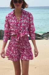 Video of a woman walking on a beach wearing a bright pink and white floral smocked mini skirt with ruffle hem, paired with a matching tied shirt, holding a straw hat and smiling.
