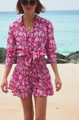 Video of a woman wearing a pink and white floral print long-sleeve shirt tied at the waist, styled with matching shorts, sunglasses, and a straw hat on the beach.