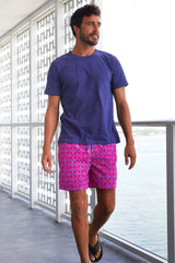 Man wearing a navy blue t-shirt and bright pink and blue patterned swim shorts, walking on a balcony in flip-flops.