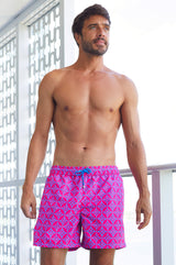 Man wearing bright pink and blue patterned swim shorts, standing on a balcony.
