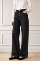 Model wearing the Aspiga Relaxed Straight Denim Jean in Black with a cream ruffle blouse tucked in, cropped front view, hand in pocket