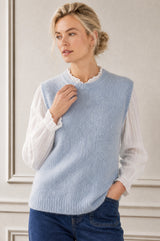 A woman wearing a light blue ribbed knit sweater vest over a white long-sleeve blouse and dark blue jeans, standing against a light beige paneled wall.