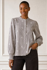 Model wearing the Aspiga Cosima Embroidered Blouse in Chocolate/White, a striped blouse with embroidered motifs and ruffled mandarin collar, smiling, hands clasped