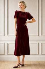 Model wearing the Aspiga Velvet Scoop Back Dress in Wine Red with short sleeves, full-length front view, hand on hip, smiling