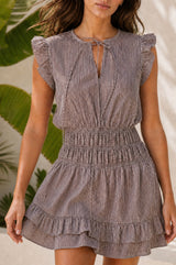 Model wearing the Aspiga Mini Macy Stripe Dress in Chocolate/White, a pinstriped mini dress with frill cap sleeves, a tie neck and a tiered ruffle hem, alternate view