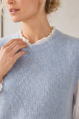 A close-up of a woman wearing a light blue ribbed knit sweater vest over a white ruffled blouse.